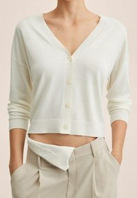 Frau trägt eine cremefarbene, kurz geschnittene V-Ausschnitt-Strickjacke mit Knöpfen und beige, hoch taillierte Hose, die Hände in den Taschen und ein umgeschlagener Hosenbund.