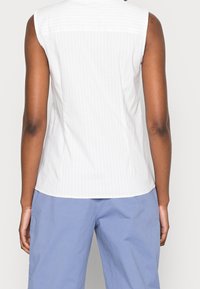 Hvid ærmeløs top med lodrette lyse striber, rund halsudskæring og buet kant. Stoffet ser glat og let ud.