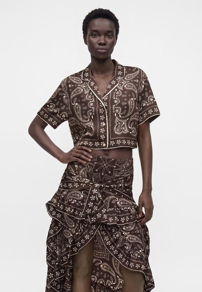 Femme portant un haut court marron à motif paisley, boutonné, et une jupe asymétrique assortie avec des détails à volants, debout, main sur la hanche.