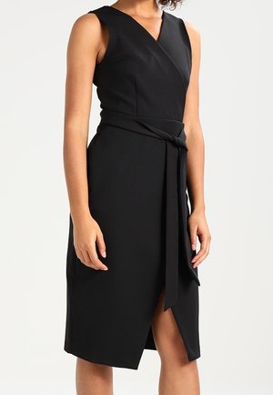 Zwart mouwloze midi-jurk met V-hals, overslag aan de voorkant, tailleband en voorsplit, gedragen door een staand persoon tegen een effen achtergrond.