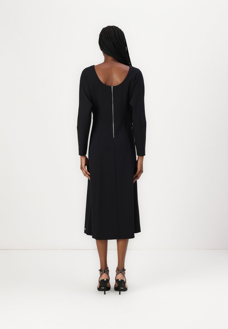 Schwarzes, langärmeliges Kleid mit rundem Rücken und Reißverschlussdetails, das am Saum leicht ausgestellt ist, getragen mit schwarzen Riemchen-Heels.