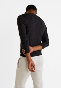 Dunkelgrauer, gerippter Pullover mit langen Ärmeln, taillierter Passform und Rundhalsauschnitt, getragen mit hellen Hosen, die Hände hinten zusammengelegt.
