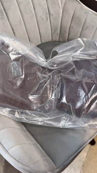 Transparante plastic tas met een donkerbruine rechthoekige object erin. De achtergrond toont een lichtgrijze gestoffeerde stoel met een gebogen ontwerp.