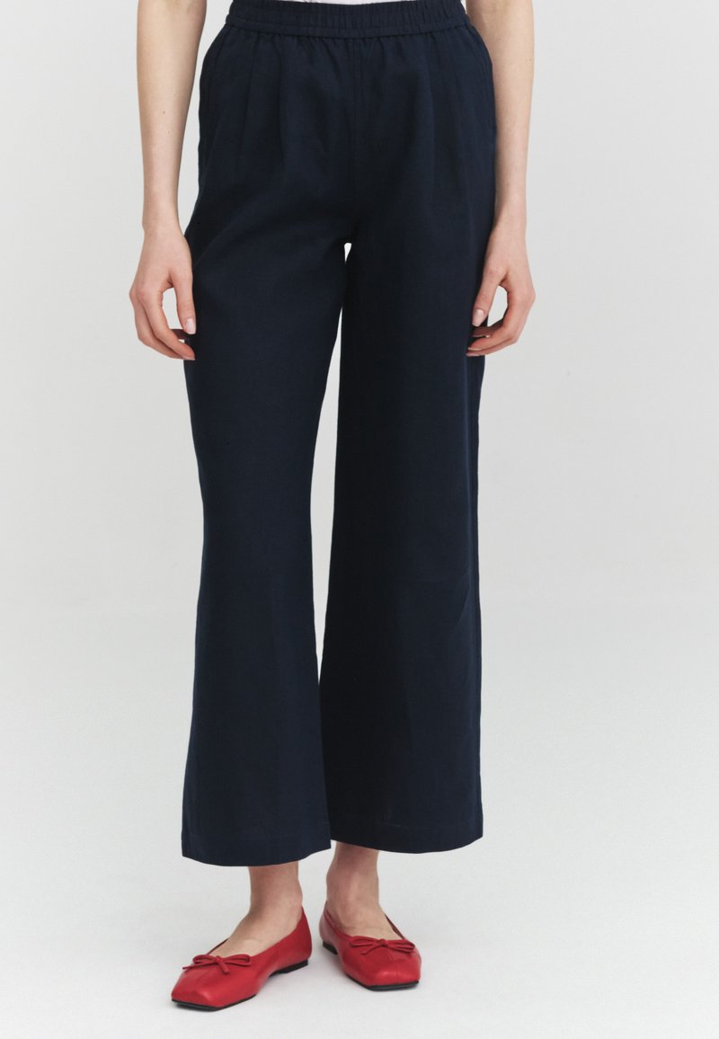 Femme portant un pantalon bleu marine ample avec une taille élastique et des ballerines rouges avec petits nœuds, debout devant un fond clair uni.