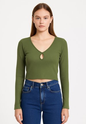 Donna con capelli castani lisci che indossa un crop top verde a maniche lunghe con scollo a goccia e jeans blu a vita alta.