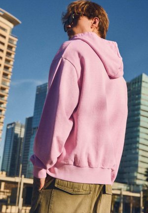 Jeune homme portant des lunettes de soleil, un sweat à capuche rose et un pantalon olive, debout les mains dans les poches dans une zone urbaine avec de hauts immeubles sous un ciel clair.