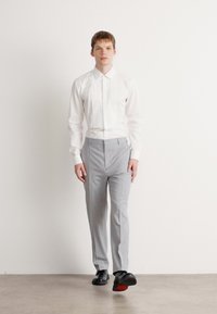 Chemise blanche à manches longues avec plis sur le devant, associée à un pantalon gris clair tailleur et des chaussures noires à semelles rouges, se tenant devant un fond neutre.