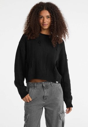 Pull cropped noir avec texture côtelée et détails distendus. Associé à un pantalon cargo gris avec une coupe décontractée et des poches latérales.