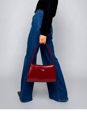 Persoon in wijde blauwe spijkerbroek en zwart topje die een kleine rode leren handtas met dubbele bandjes vasthoudt, staand tegen een effen grijze achtergrond.