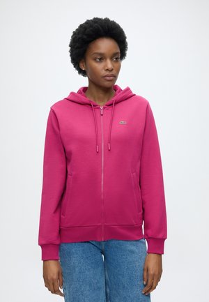 Femme aux cheveux courts et bouclés, portant un sweat à capuche zippé rose vif et un jean bleu, debout devant un fond blanc uni.