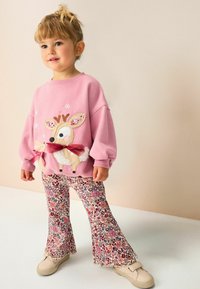 Sweatshirt roz cu aplicație de ren și fundiță, combinat cu pantaloni evazați florali în nuanțe de roșu, roz și crem. Pantofi bej.
