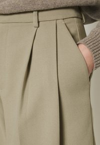Beige high-waisted broek met plooien en zijzakken, gemaakt van een textuurstof. Kenmerkt zich door strakke lijnen en minimale hardware.