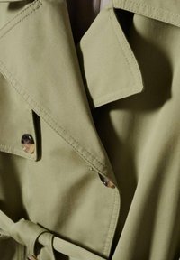 Gros plan d’un trench-coat kaki montrant son large col, bouton et détail de ceinture avec coutures visibles.