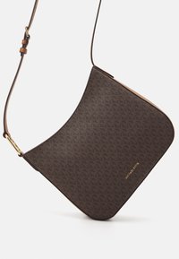Bolso bandolera marrón con un diseño estampado, forma curva, correa de cuero y adornos en dorado, con el nombre de la marca en la parte frontal.