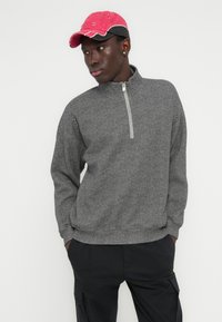 Sweat-shirt gris en chevron avec un col zippé quarter zip, manches longues et coupe décontractée. Associé à une casquette de baseball rose et noire.