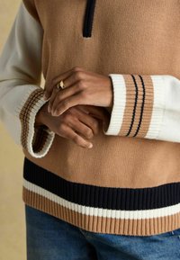 Prindi õhuke kudum pruunis ja koorevärvis, kontrastsete soonikservadega. Omab pool-zip kaelust ja triibulisi aktsente varrukate otsas.