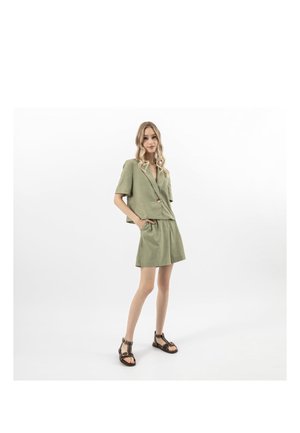 Donna in piedi che indossa un blazer verde oliva chiaro a maniche corte e gonna abbinata, con sandali neri borchiati, su sfondo bianco uniforme.