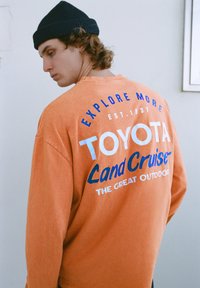 Oranžová košeľa s dlhými rukávmi s modrým a bielym grafickým textom na chrbte, s textúrovanou látkou a nadrozmerným strihom.