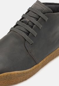 Zapatilla de cuero gris oscuro con un acabado suave, que presenta cordones grises planos, un diseño cosido y una suela de goma texturizada.