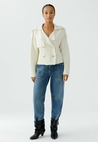 Frau, die einen cremefarbenen gestrickten, doppelt geknöpften Cardigan, blaue hochtaillierte Jeans mit aufgesetzten Taschen und schwarze, lässige Stiefeletten trägt.