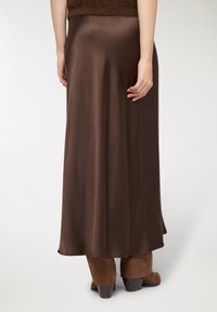 Gonna lunga in satin marrone con una texture liscia, caratterizzata da una silhouette svasata e un leggero effetto scintillante, abbinata a stivali marroni con tacco.