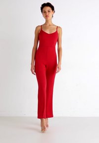Combinaison rouge avec des bretelles fines, un corsage ajusté et des jambes évasées. Confectionnée en tissu extensible pour le confort. Design clair et minimaliste sans motifs.