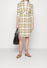Abito camicia con bottoni in fantasia quadretti, caratterizzato da tonalità gialle e bianche, stretto in vita con una cintura bianca. Gli accessori includono una borsa nera e sandali marroni.