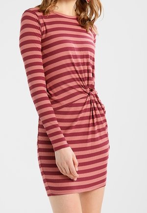 Kvinde iført en langærmet, pink og burgunder vandret striped kjole med en samlet knudedetalje i taljen.