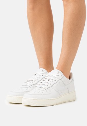 Piernas que llevan zapatillas Nike Air Force 1 blancas con cordones, de pie sobre un fondo blanco.