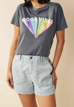 Kvinde iført en falmet mørkegrå t-shirt med farverig "Good Vibes" tekst og lyse denimbukser, stående med hænderne i lommerne.