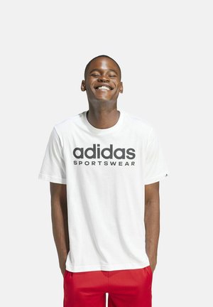 Maglietta in cotone bianco con un grande logo nero "adidas SPORTSWEAR", collo rotondo e maniche corte, abbinata a pantaloni corti rossi.