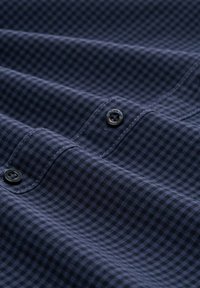 Tessuto in blu navy a quadretti con una sottile texture e due bottoni neri visibili, che mettono in evidenza una trama liscia e un design strutturato.