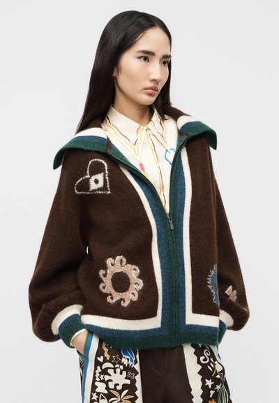 Νεαρή γυναίκα που φοράει καφέ πουλόβερ με φερμουάρ, διακοσμημένο με αφηρημένα σχέδια καρδιάς και σπειρών, συνδυασμένο με παντελόνι με σχέδια και πουκάμισο με γιακά.