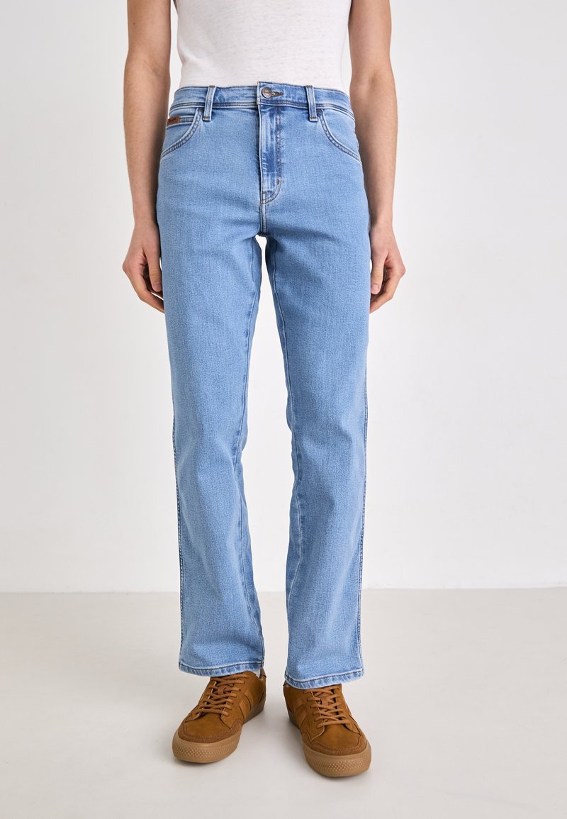 Vaqueros de mezclilla azul claro con corte recto, diseño de cinco bolsillos y cierre de botón de metal. Combinados con zapatillas de ante marrón.