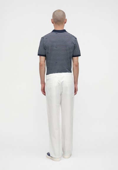 Homme au crâne rasé portant un polo rayé bleu marine et blanc, un pantalon blanc et des baskets foncées, debout de dos sur un fond uni.