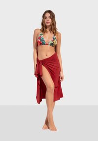 Bikini dengan atasan bermotif bunga dan rok lilit merah solid yang memiliki detail ikatan dan belahan tinggi, terbuat dari kain yang halus dan ringan.