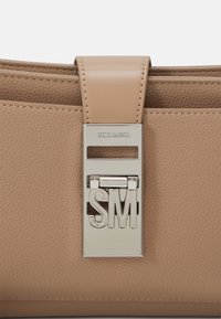 Sac à main en cuir texturé beige avec une fermeture argentée présentant les lettres découpées "SM" et "Steve Madden" gravées au-dessus.