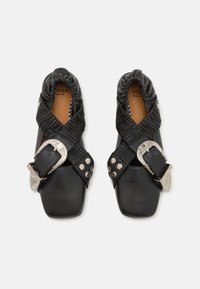 Zapatos de cuero negro con punta cuadrada, diseño de tiras elásticas entrelazadas, hebilla plateada y detalles de tachuelas. Parte superior de textura suave y suela plana.