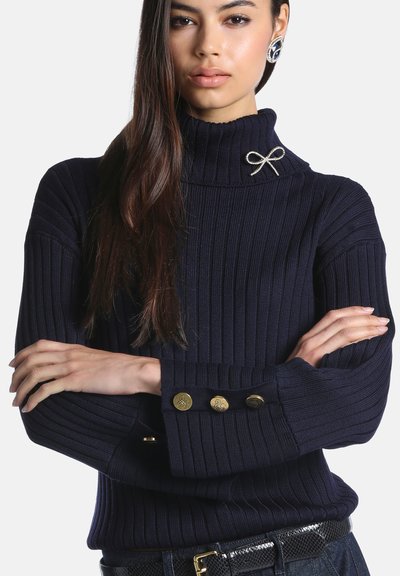 Femme aux longs cheveux bruns porte un pull à col roulé côtelé bleu marine avec des boutons dorés et une broche décorative en forme de nœud sur le col.