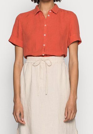 Femme portant une chemise orange à manches courtes boutonnée, rentrée dans une jupe beige taille haute avec un nœud coulissant à la taille.