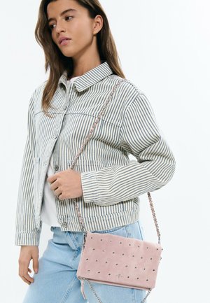 Young woman wearing a striped jacket and light blue jeans, carrying a pink studded shoulder bag with a chain strap.