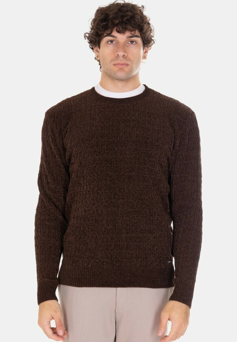 Giovane con capelli ricci che indossa un maglione lavorato a trecce di colore marrone scuro e pantaloni beige chiaro contro uno sfondo chiaro uniforme.