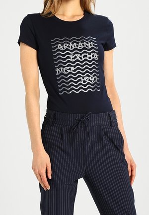 Laivansinis t-paita lyhyillä hihoilla, jossa on hopeinen aalto-kuvio ja teksti "ARMANI EXCHA NICE 1991", yhdistettynä raidallisiin laivansinsisiin housuihin.