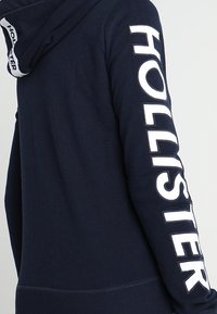 Felpa blu navy con un grande logo bianco "HOLLISTER" lungo la manica sinistra, dotata di cappuccio con cordino e orlo a coste. Tessuto morbido.