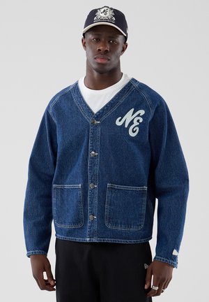 Mand iført blå denim-knapjakke med "NE" logo på brystet, hvid t-shirt, sorte bukser og navy baseballkasket med logo.