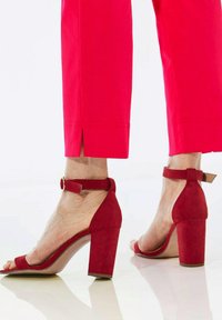 Sandales à talons en daim rouge avec un talon fermé, une pointe ouverte et une sangle autour de la cheville. Le talon carré offre de la stabilité ; finition lisse et texturée.