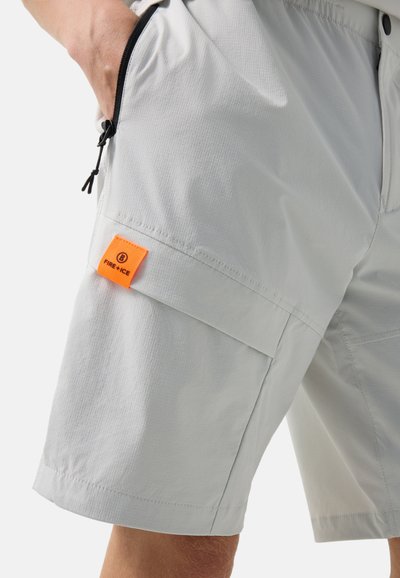 Zbliżenie jasnoszortów z zamykaną na suwak kieszenią i pomarańczową metką marki "FIRE + ICE" na udzie, ręka spoczywa w bocznej kieszeni.