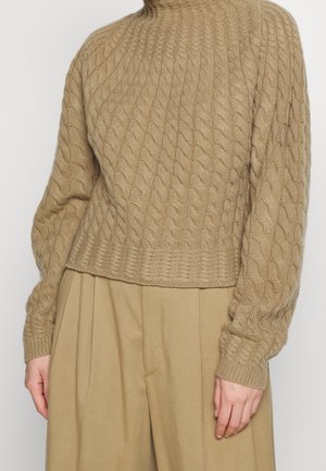 Beige kabelstrikket sweater med høj hals, der har ribbede ærmer og kant, parret med lyse plisserede bukser.