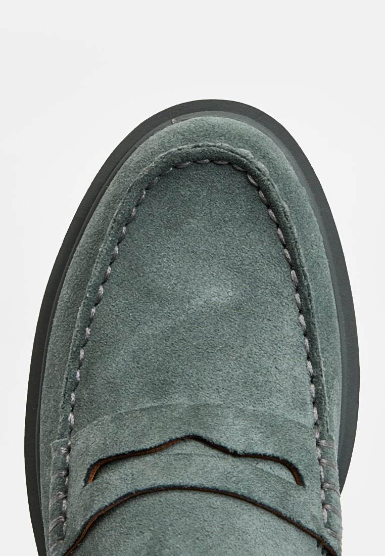 Mocassin en suede vert avec une texture lisse, présentant des coutures ton sur ton et un design classique, mettant en avant l'embout arrondi et la semelle en caoutchouc.