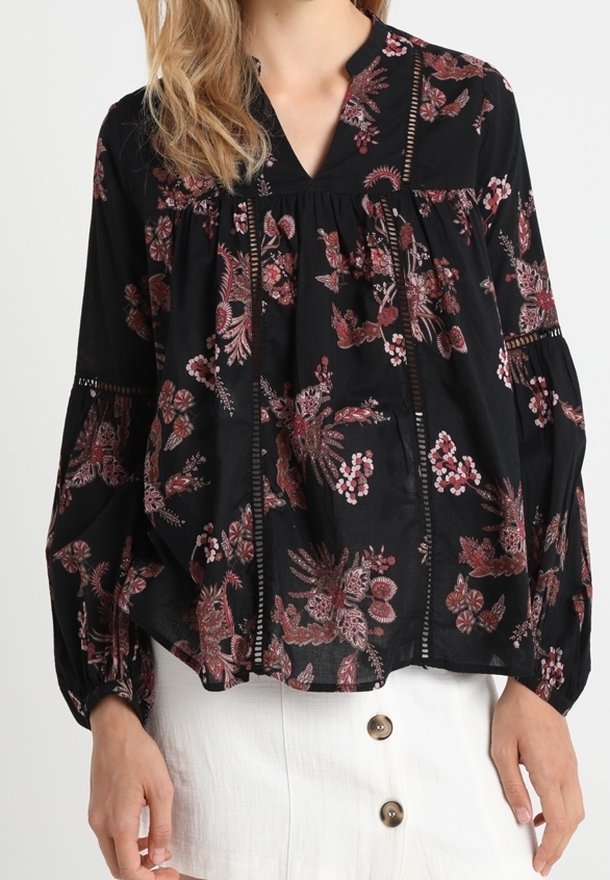 Blouse noire à manches longues avec motifs floraux roses, inserts en dentelle et col en V, associée à une jupe blanche à deux boutons.